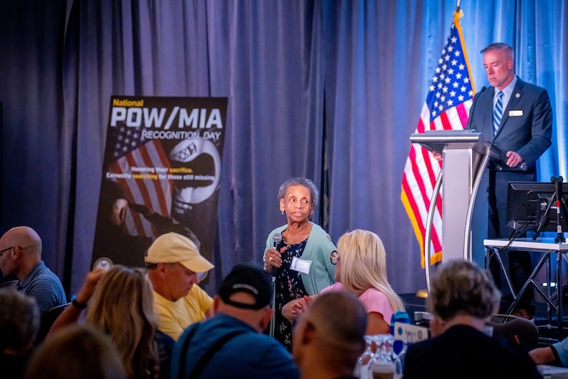 A person speaks in front of a large group of people in a banquet hall as another person stands at a podium in the background.