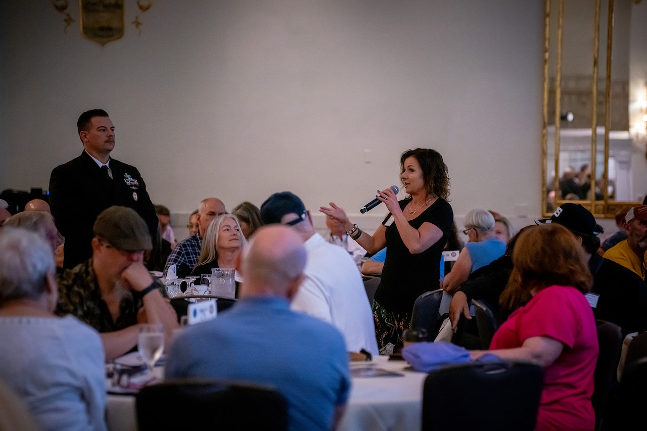A person with a microphone stands to talk to a crowd of people seated at tables.