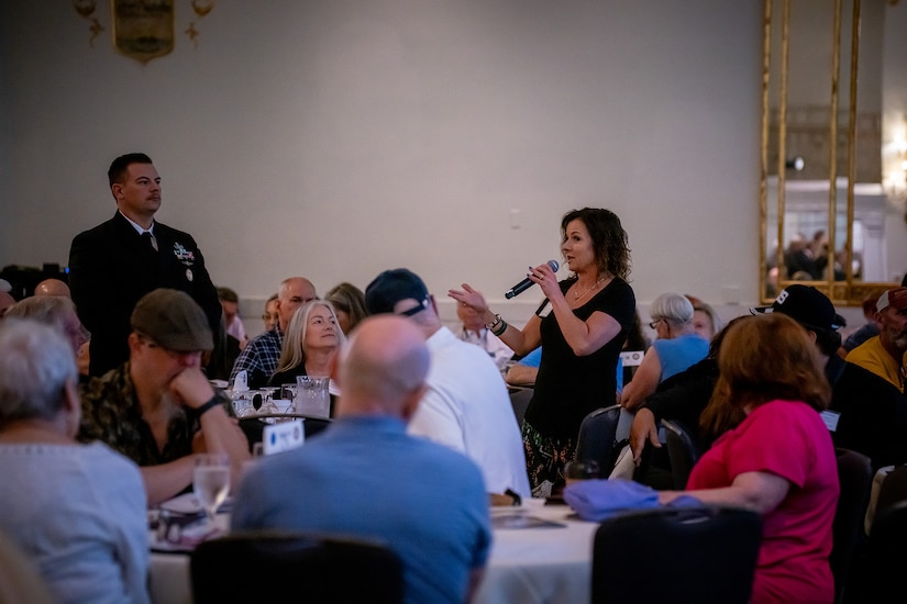 A person with a microphone stands to talk to a crowd of people seated at tables.