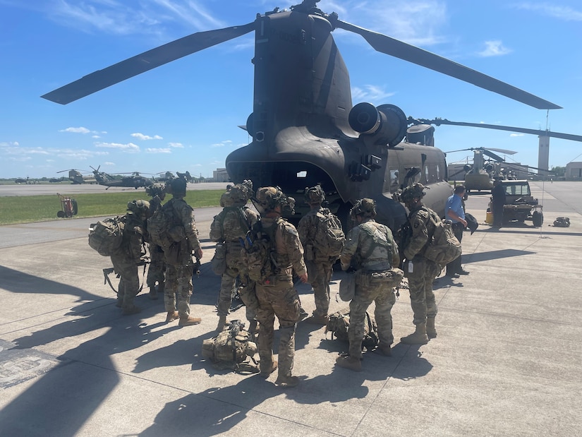 Eight soldiers stand behind a parked helicopter on a sunny day on the tarmac.