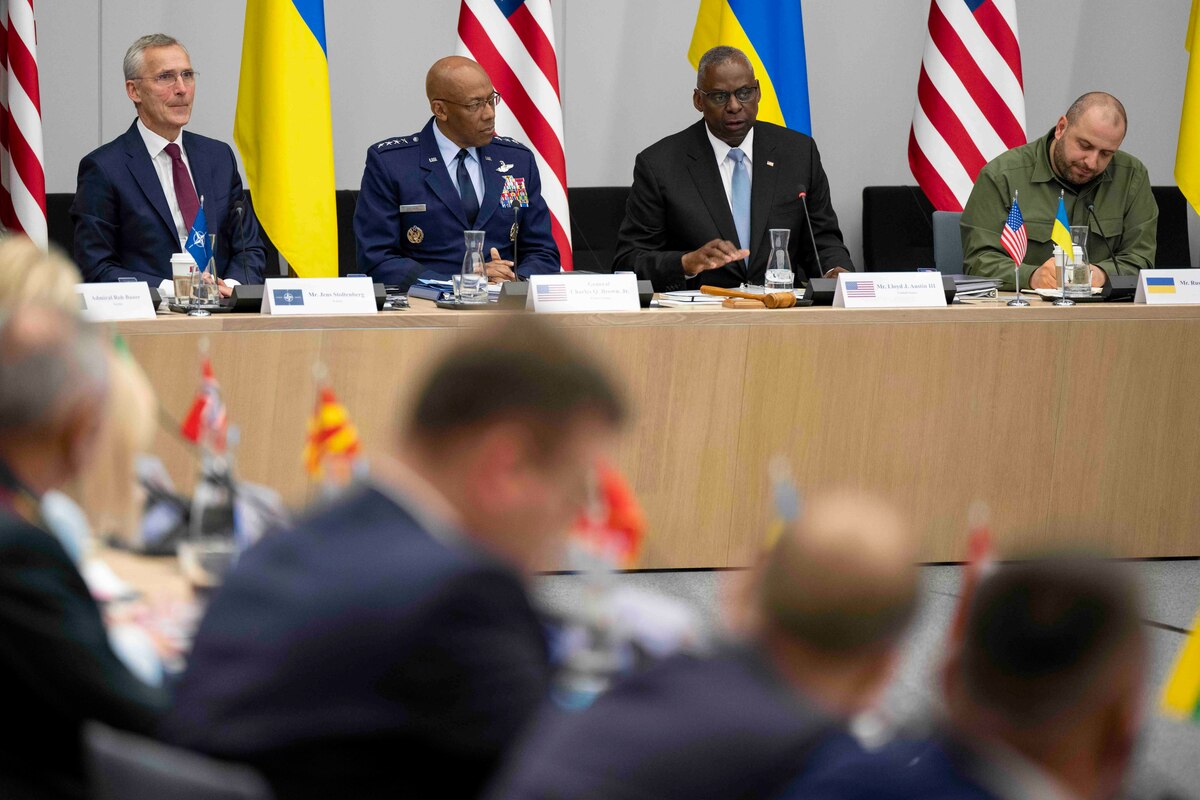 Two men wearing civilian attire and two men wearing military uniforms sit at a table displaying U.S. and Ukrainian flags.