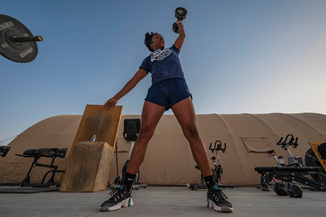 An airman lifts a weight over their head in front of exercise machines with brown tent in the background.