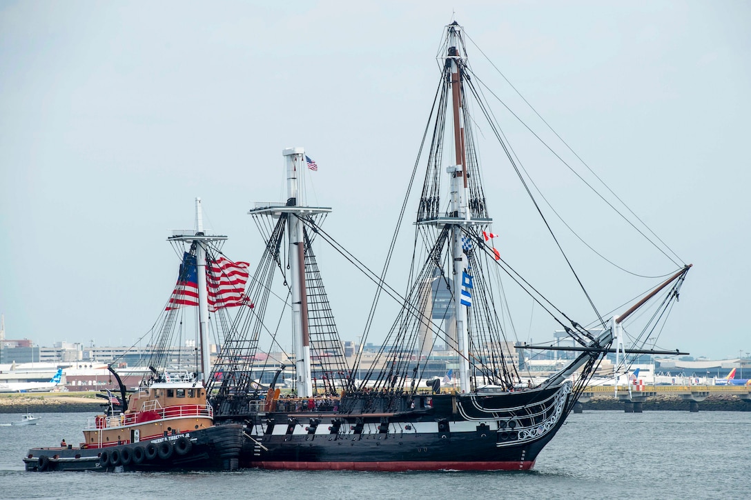 A boat rides next to a three-masted ship in a body of water with a city’s skyline in the background.