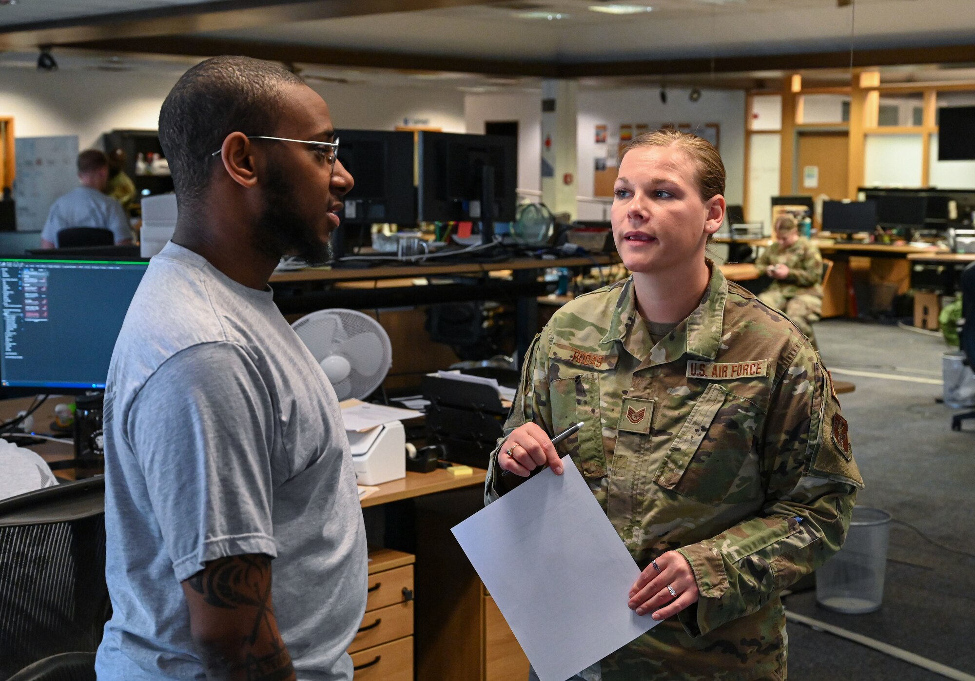 Two Airman, one in a while shirt and one in a green Air Force uniform talk while holding papers.