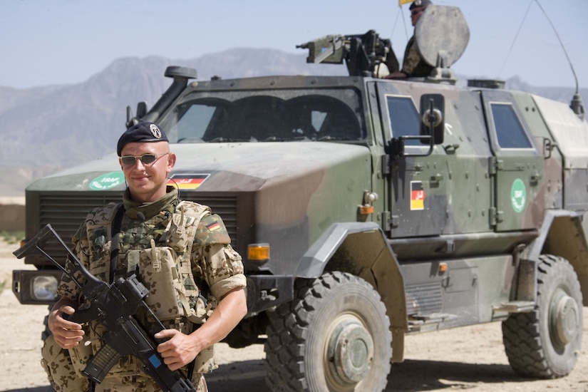 A man in uniform, holding an automatic weapon, stands in front of an armored vehicle.