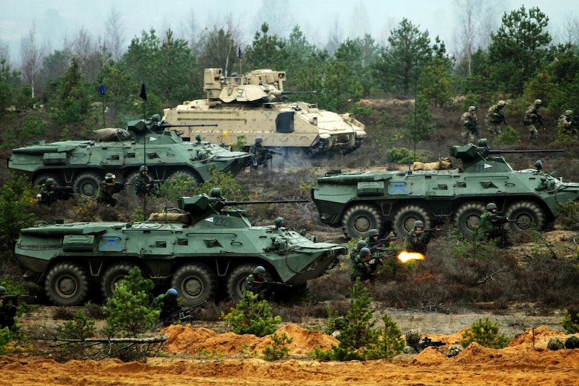 Armored vehicles transverse a field as soldiers, on foot, complete the exercise.