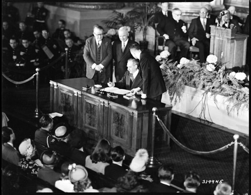 Three men look on as a fourth signs a document during a large ceremony.