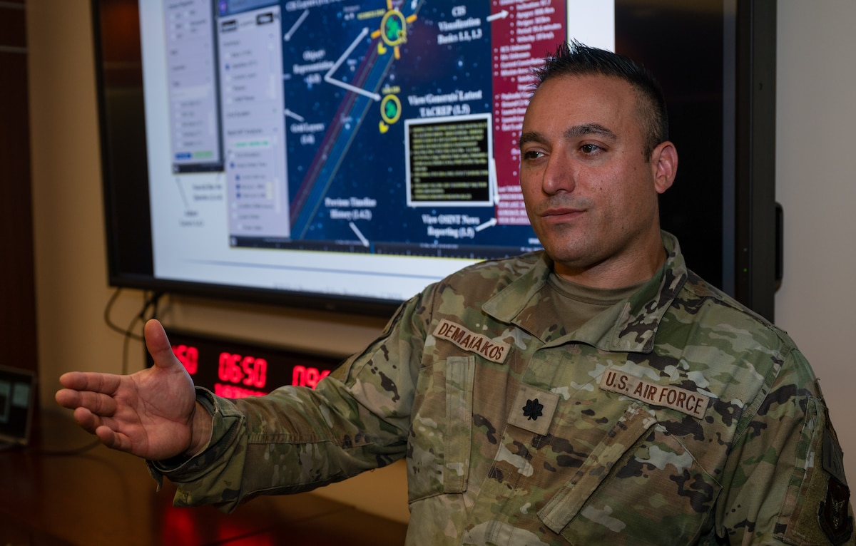 A man wearing a U.S. Air Force uniform stands in front of a computer monitor displaying a graphic.