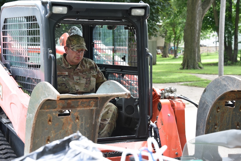An airman operates a medium-sized motorized cleanup vehicle with trees in the background.