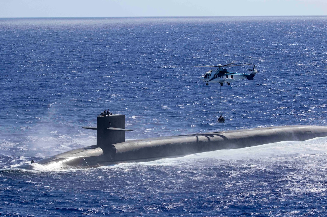 A helicopter lowers a container by cable to a submarine in the ocean.