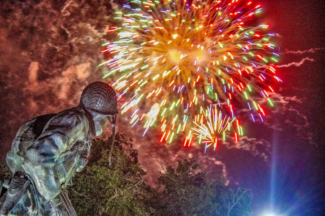 Fireworks explode in a bright smoky sky above a statue and trees at night.