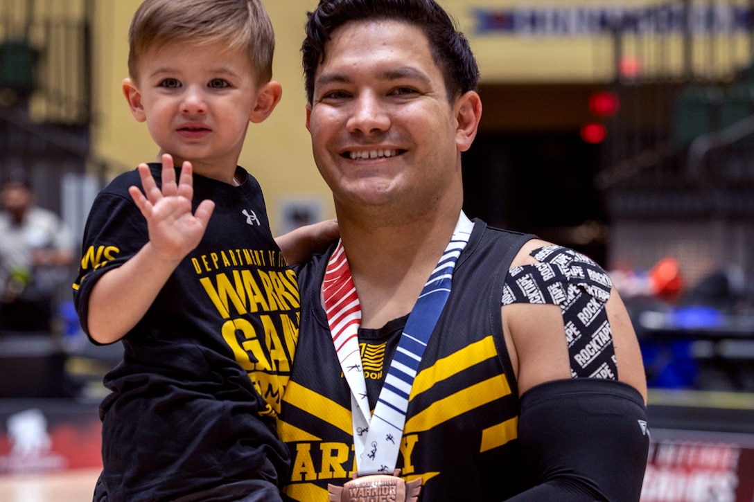 A soldier wearing a medal smiles in a gym while holding a child, who waves at the camera.