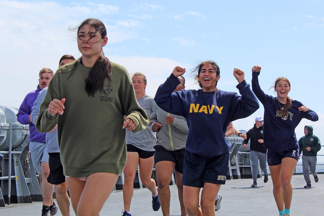 Two sailors raise their arms during a daylight group run on the deck of a Navy ship.