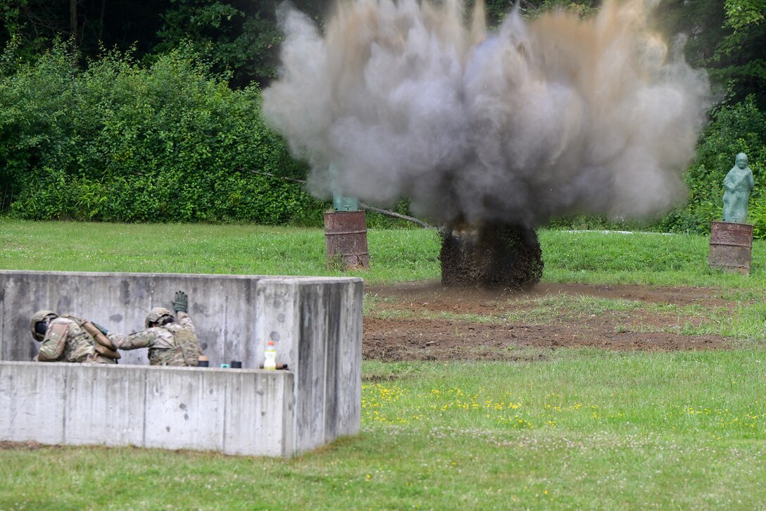 Two airmen take cover behind a cement wall as a grenade detonates in the grassy area, creating smoke and dust.