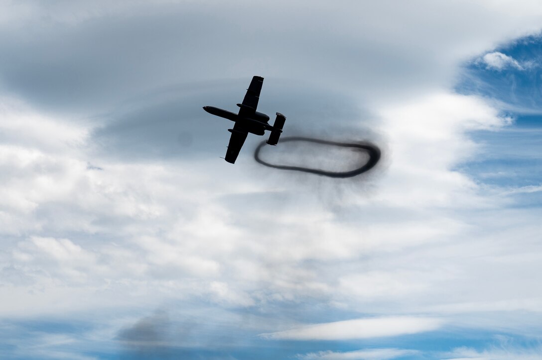 Photo shot from below shows a military aircraft creating a dark oval contrail against a mostly cloudy sky.