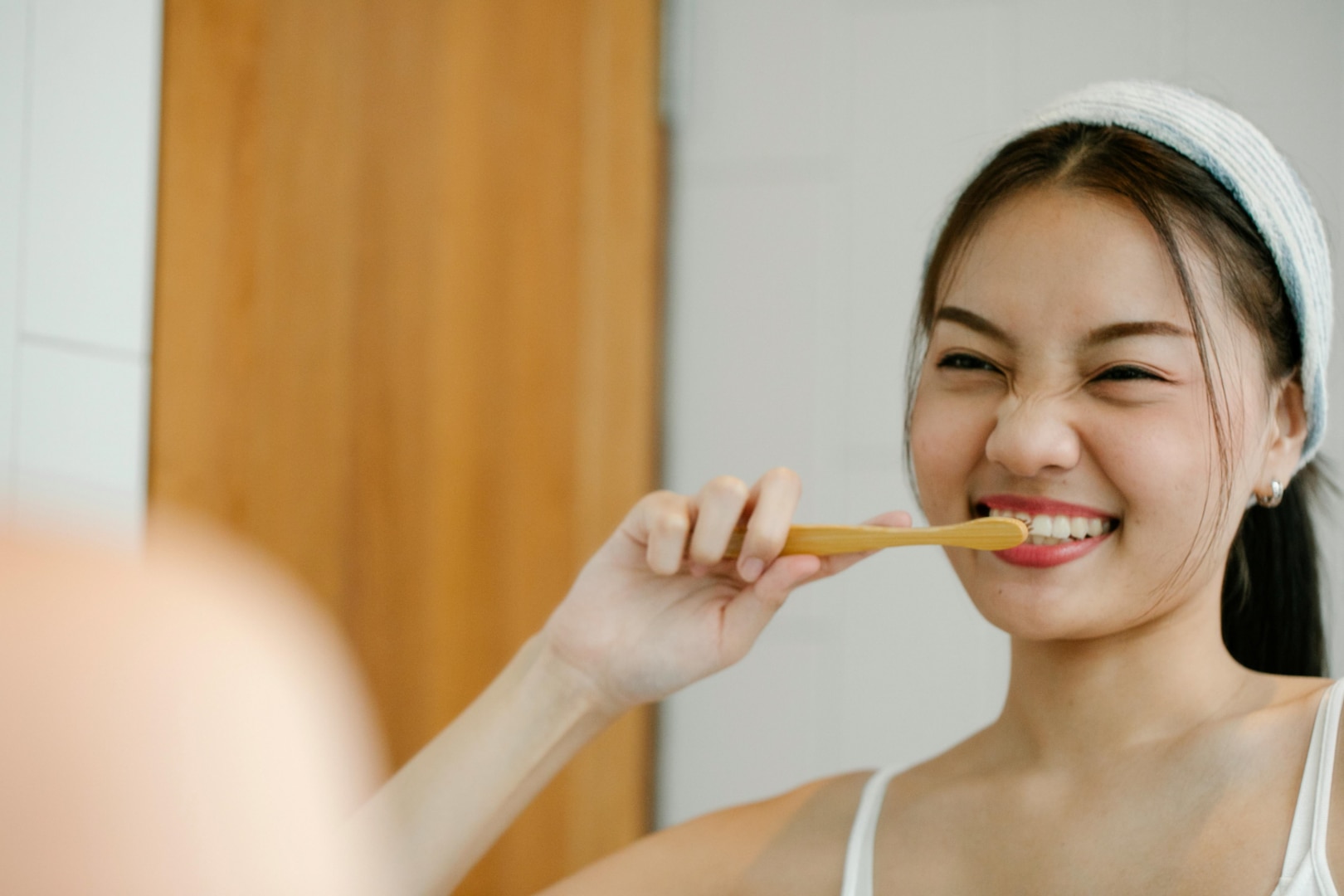 Woman wearing white headband looks in mirror while brushing teeth.