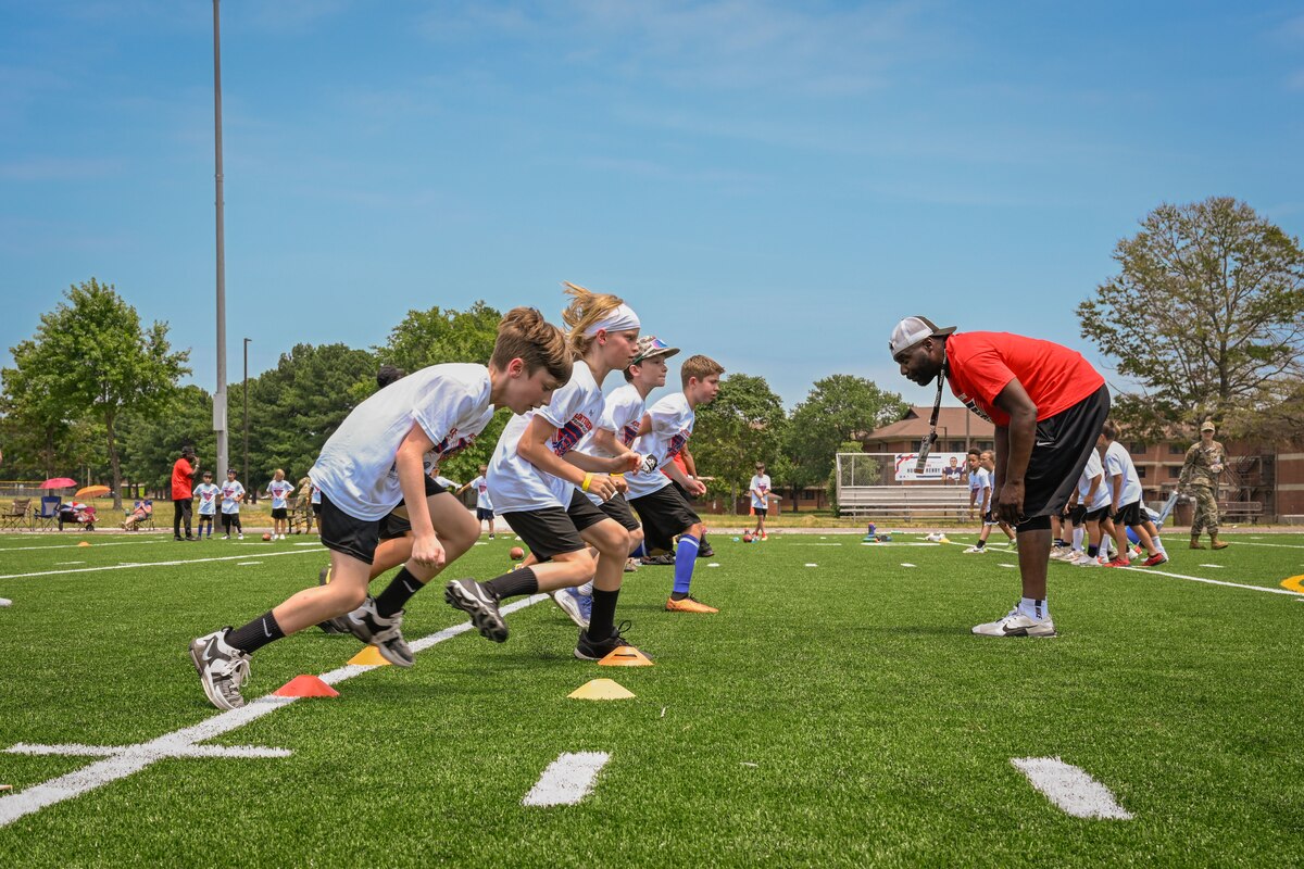 A coach wearing a red shirt and backwards hat stands to the right of a line of children wearing white shirts who participate in drills during a football camp.
