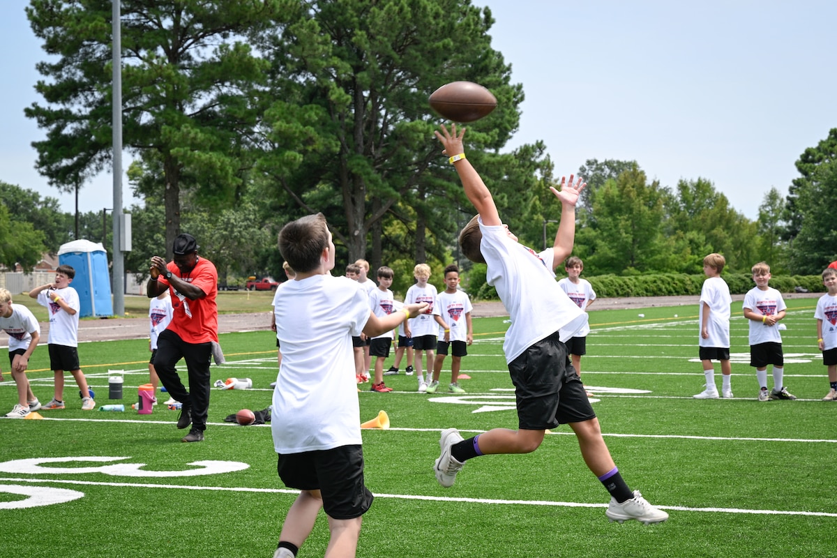 A boy wearing a white shirt and black shorts catches a football with one hand during a football camp.