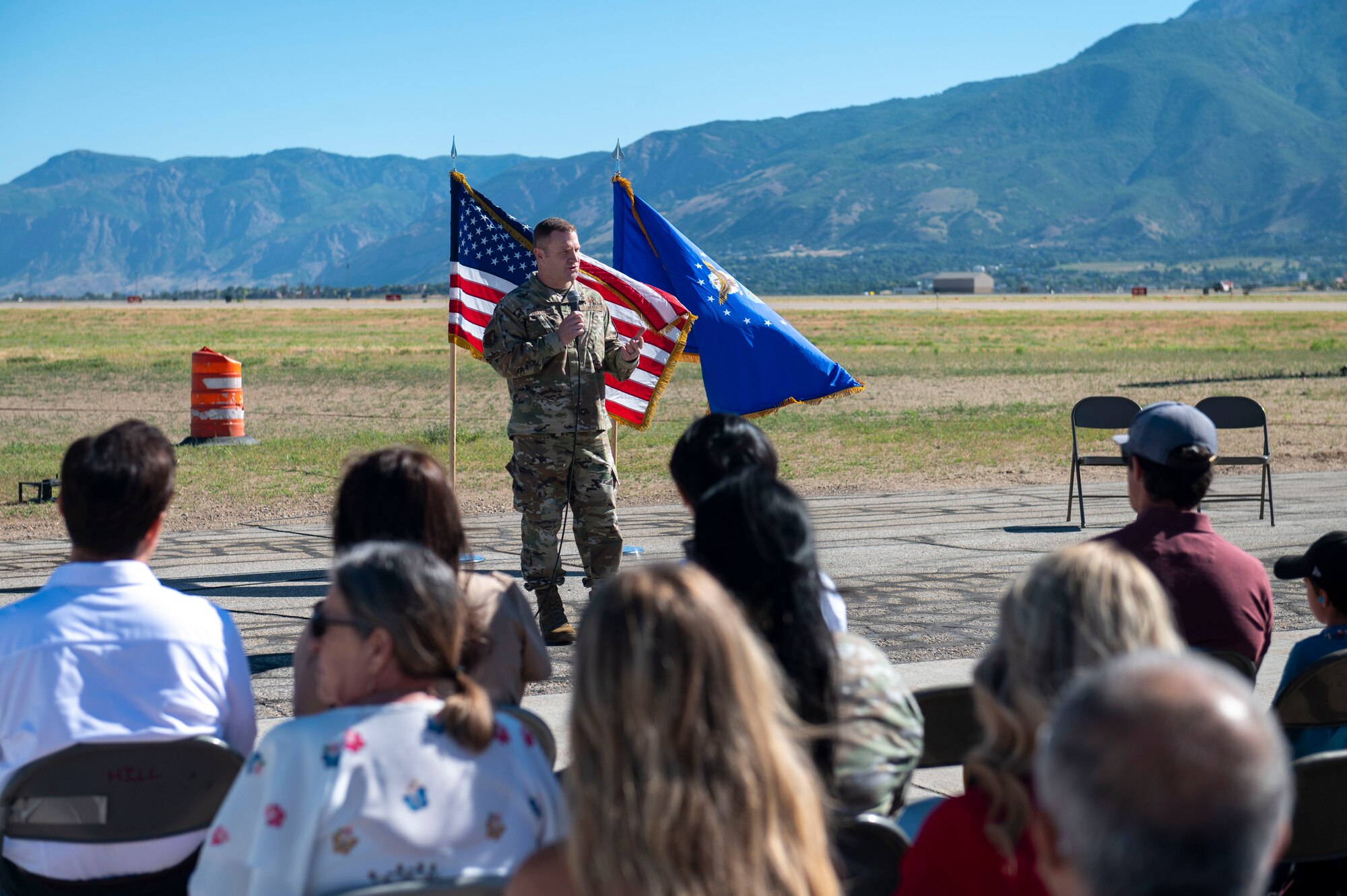 Col. Holland stands speaking to seated guests on the airfield.