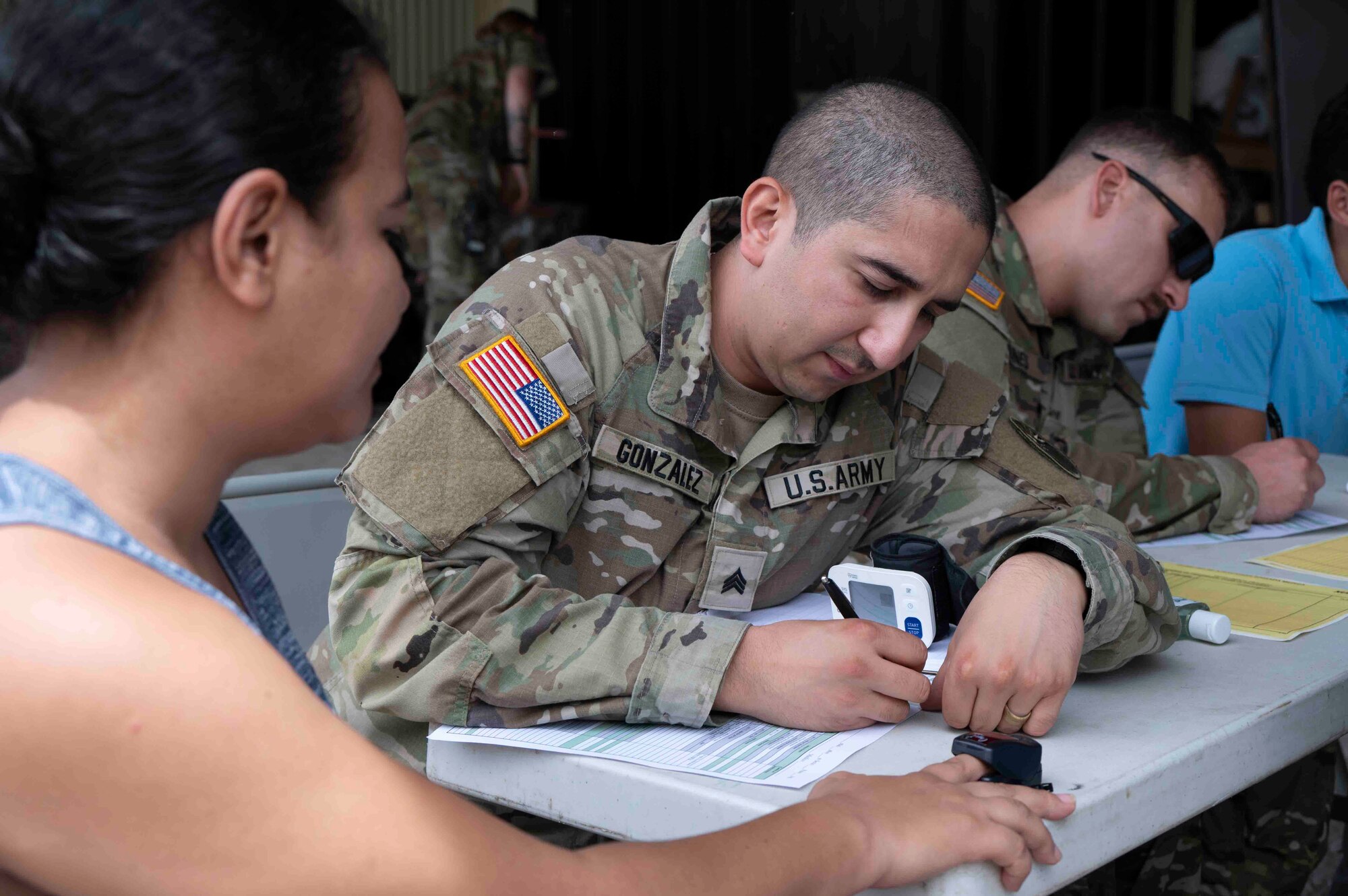 A photo of a military medical professional writing on a form while a patient has a blood pressure clip on.