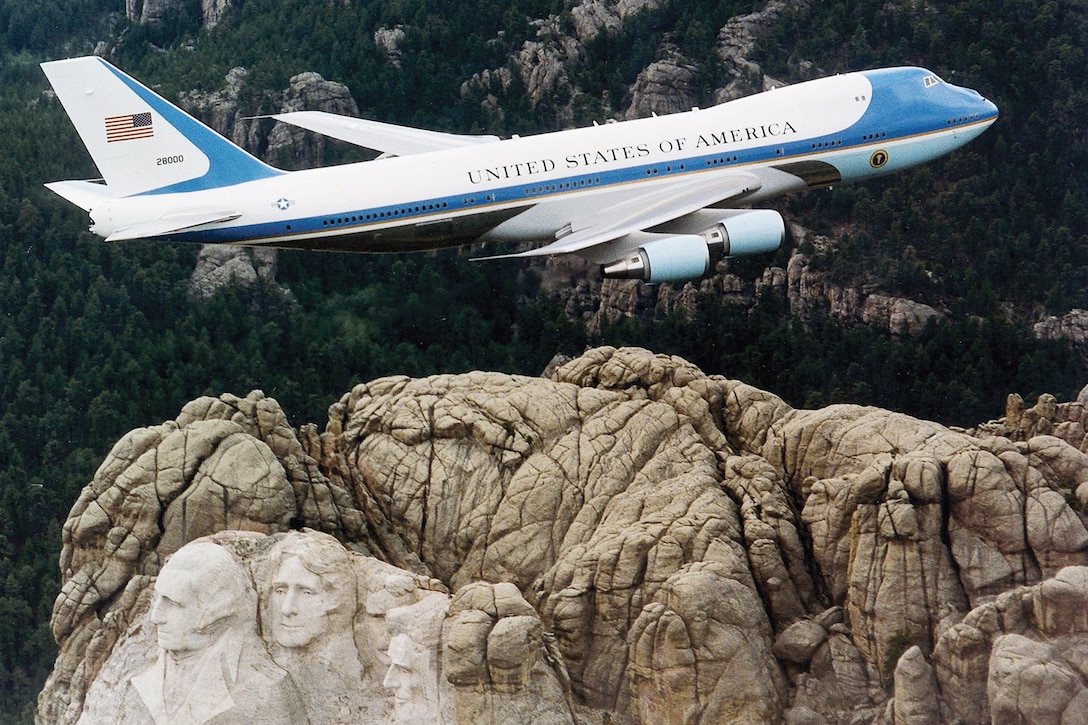 An aircraft flies over a monument of presidents carved at the top of a mountain.