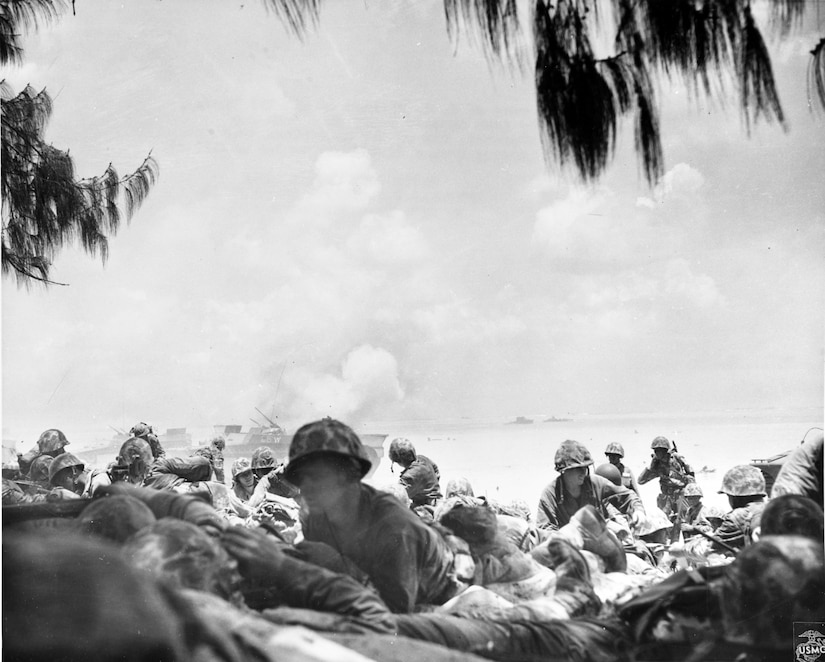 Several men lie prone on a beach. Smoke can be seen in the background.