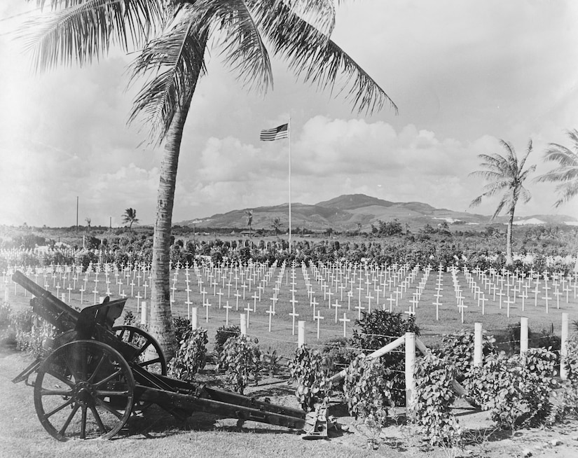 Rows of white crosses line a graveyard. A U.S. flag is waving in the background while a cannon rests in the foreground.