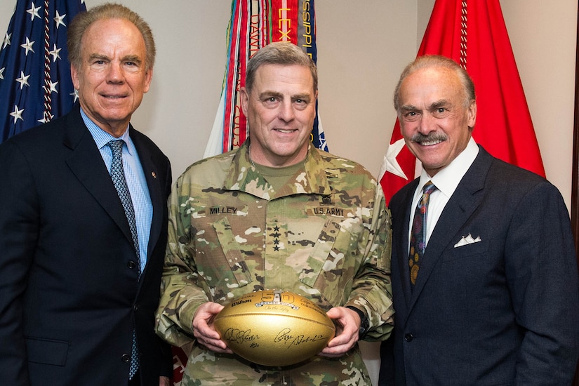 A person in a military uniform stands between two others in civilian clothing. The service member is holding a football.