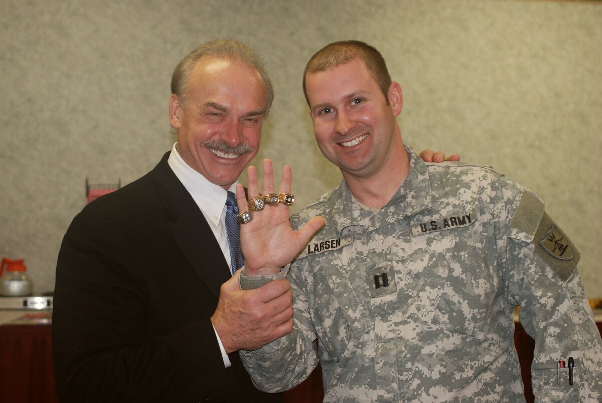 A soldier holds up their hand displaying four Super Bowl rings, as a civilian smiles and stands with their hand on the soldier's shoulder.