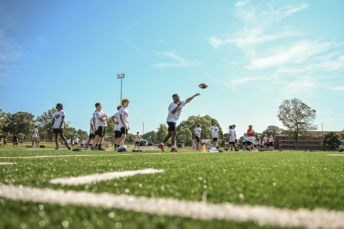 Children stand in a line as one throws a pass on a turf field during a football camp.