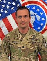 man wearing U.S. Army uniform standing in front of two flags.