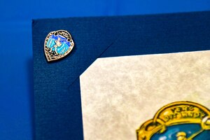 The silvered covered Military Marching Badge skills badge is presented as part of the Norwegian Ruck March ceremony at the Powell Event Center, Goodfellow Air Force Base, Texas June 27, 2024. To earn a Norwegian Armed Forces skills badge, participants must finish the course within a specific time limit determined by their age and gender. (U.S. Air Force photo by Airman 1st Class James Salellas)