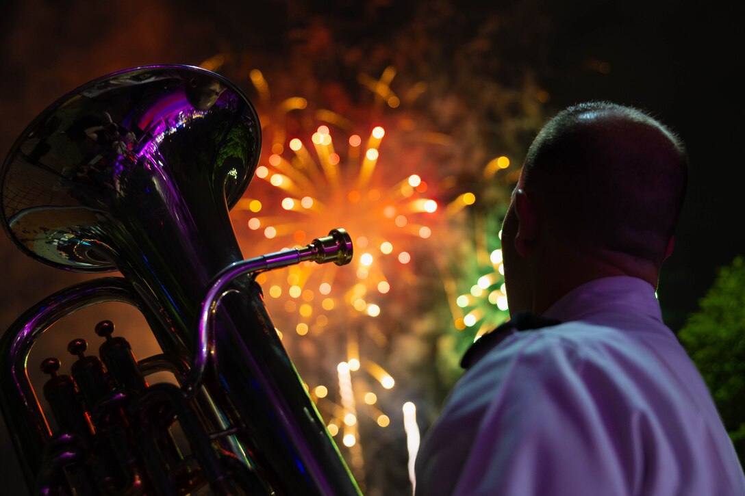 A soldier holds a euphonium while fireworks go off in the background.