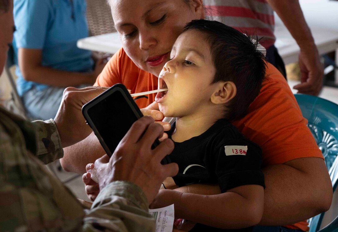A photo of a military medical professional using a wooden stick and a cell phone flashlight to see inside a child's mouth while his mother holds him.