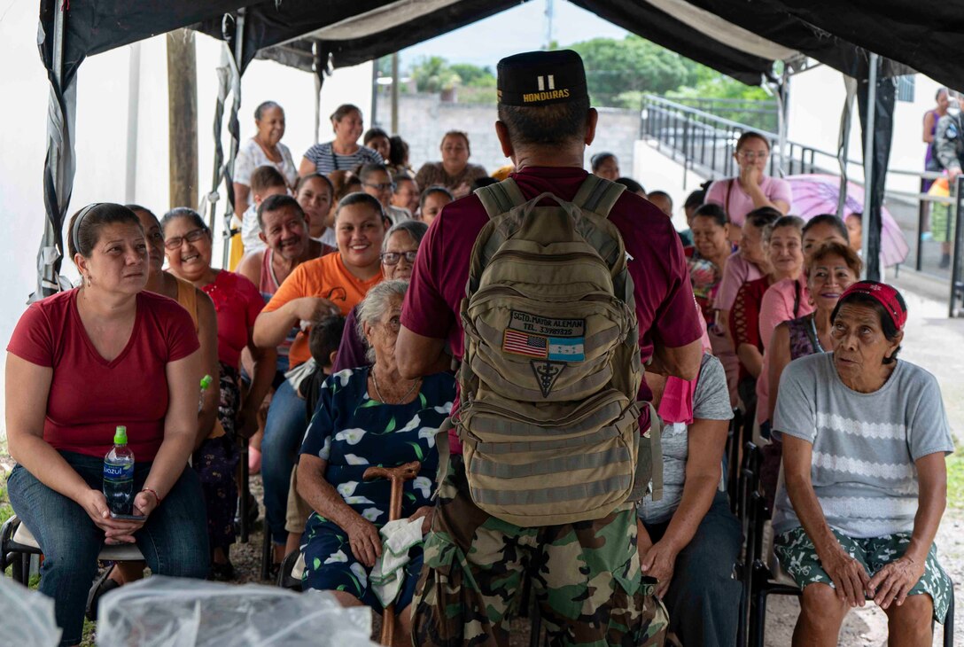 A photo of a person standing in front of a group of waiting patients talking to them.
