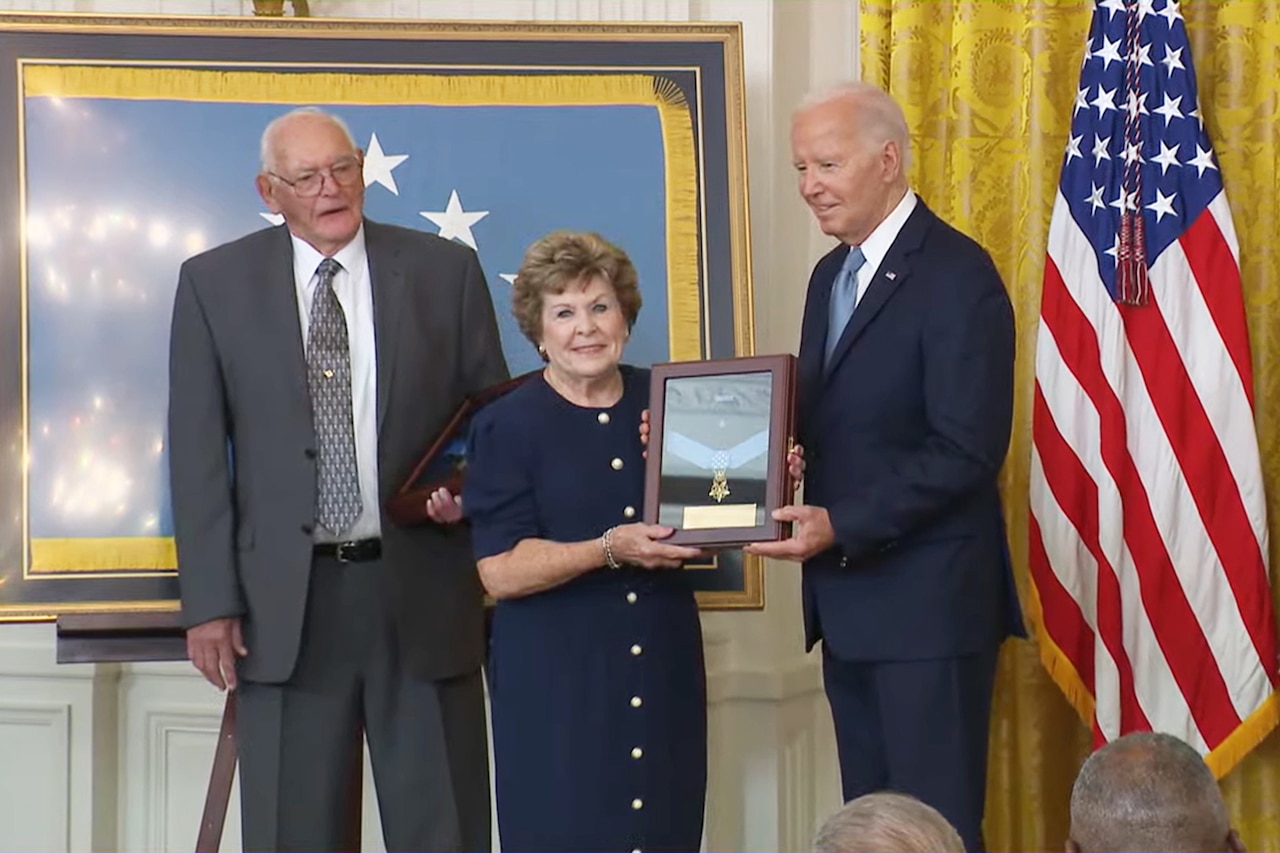 Three people stand on a stage with a U.S. flag on the far right. One holds a frame containing a medal.