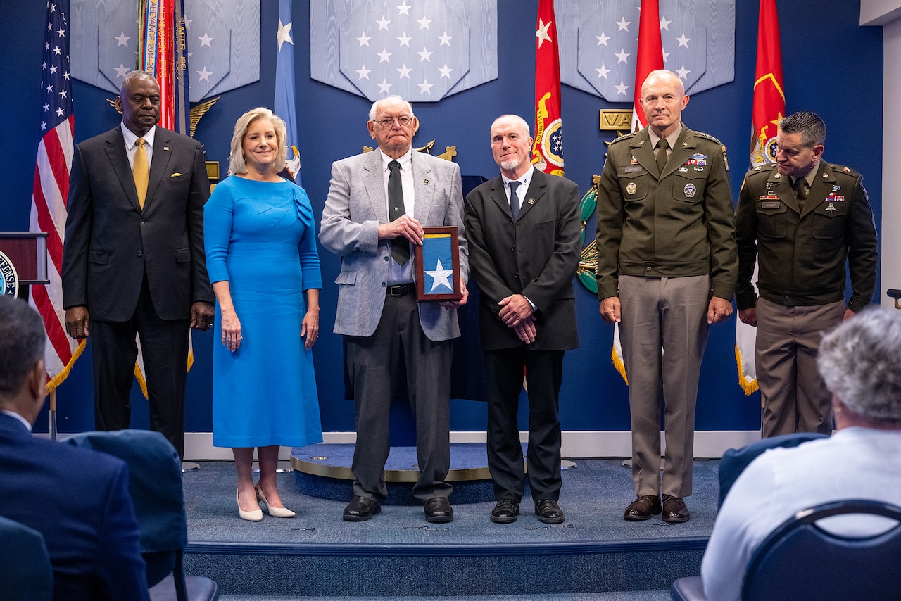 Six people stand on a stage. A person in the center holds a frame with a folded flag inside.