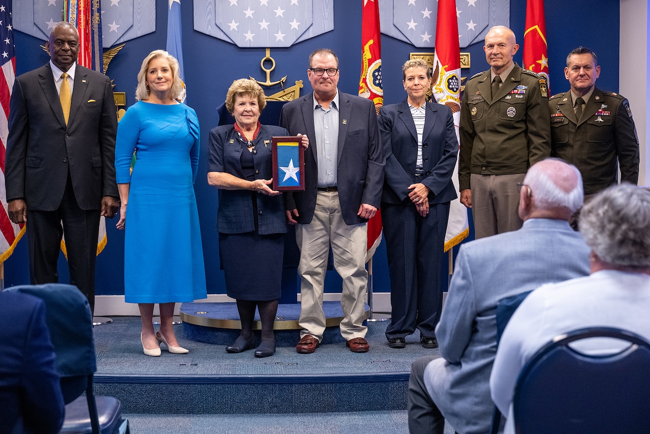 Seven people stand on a stage. A person in the center holds a frame with a folded flag inside.