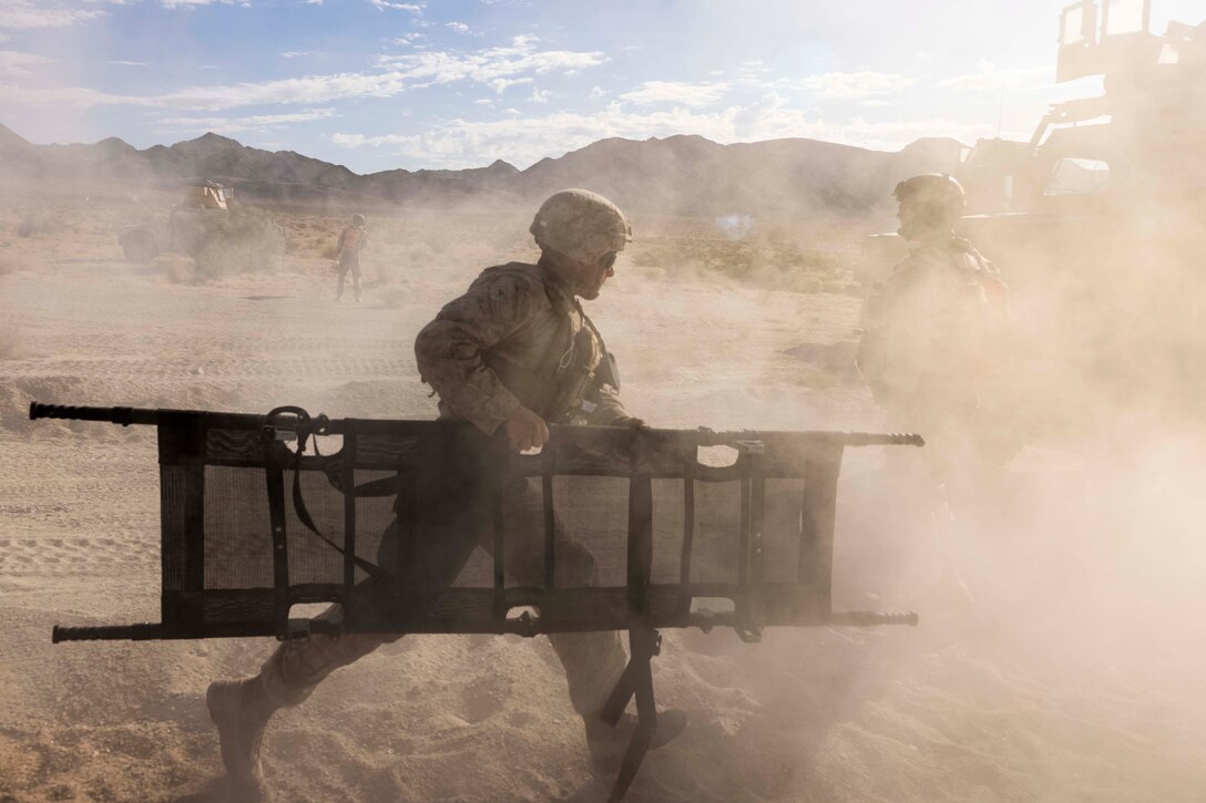 A Marine carries a litter while running through a sandy area as dust blows through the air.