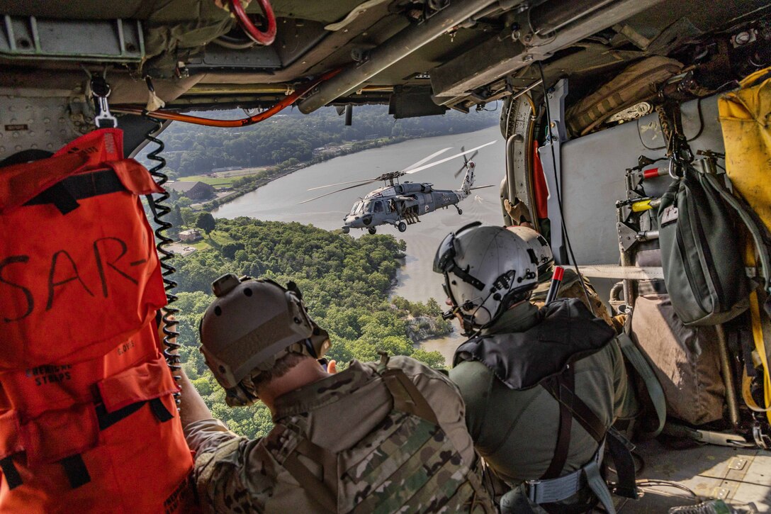 Two sailors sit in a helicopter and look out over a park and lake while another helicopter flies close by.