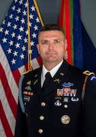 Army Soldier in dark service uniform with numerous ribbons and badges affixed. Behind him are the US flag and Army flag with battle ribbons.