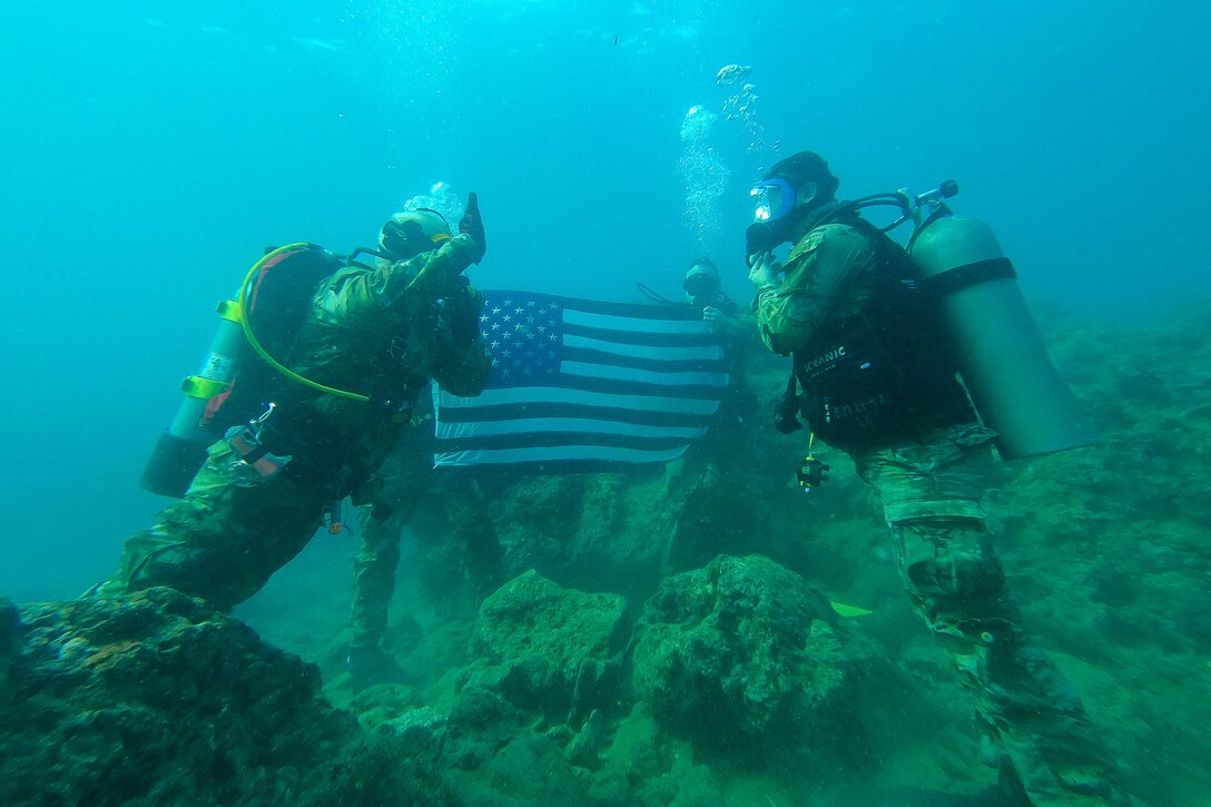 Soldiers underwater using diving gear face each other while raising their right hands as two fellow soldiers hold an American flag in the background.