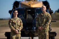 Two soldiers pose in front of an Army VH-60M "gold top" Black Hawk helicopter. They are wearing Army camouflage uniforms with no hats.