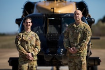 Two soldiers pose in front of an Army VH-60M "gold top" Black Hawk helicopter. They are wearing Army camouflage uniforms with no hats.