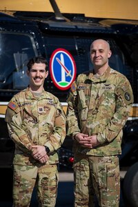 Two soldiers post next to an Army VH-60M "gold top" Black Hawk helicopter. The Military District of Washington logo on the helicopter is visible between the two soldiers.