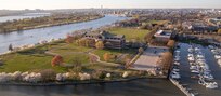 Overhead view of Ft. McNair, with large green lawns surrounding a large, red brick building in the foreground and a marina filled with boats on the right.
