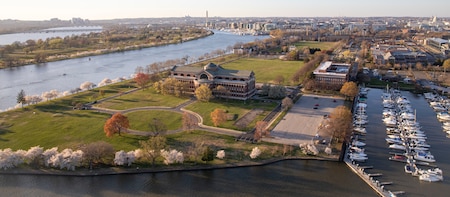 Overhead view of Ft. McNair, with large green lawns surrounding a large, red brick building in the foreground and a marina filled with boats on the right.