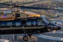 An Army VH-60M "gold top" Black Hawk helicopter flies over the Washington, D.C. area buildings. The top half of the helicopter is painted gold, and the bottom half is dark green with a gold, horizontal stripe down the middle.