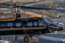 An Army VH-60M "gold top" Black Hawk helicopter flies over the Washington, D.C. area buildings. The top half of the helicopter is painted gold, and the bottom half is dark green with a gold, horizontal stripe down the middle.