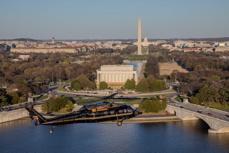 An Army VH-60M "gold top" Black Hawk helicopter flies over the Potomac near the Lincoln Memorial with the Washington Monument and U.S. Capitol building in the distance.